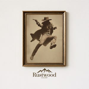 May include: Sepia-toned framed print of a cowboy in mid-air, aiming a pistol. The cowboy is wearing a hat, coat, and boots. The print is in a gold-colored frame. The bottom of the frame has the text "Rustwood GALLERY".