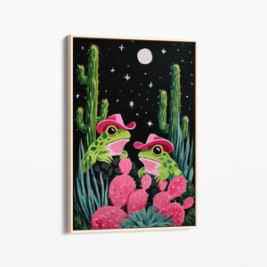 May include: A framed painting depicts two green frogs wearing pink cowboy hats, set against a night sky with a full moon and stars. The frogs are surrounded by cacti and pink prickly pear cactus. The artwork has a whimsical, illustrative style.