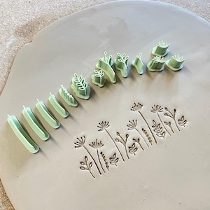 Könnte beinhalten: Eine Sammlung hellgrüner Tonstempel aus Kunststoff, die in einer Reihe angeordnet sind und verschiedene botanische Formen aufweisen. Unter den Stempeln zeigt ein flaches Stück grauer Ton eine Reihe eingeprägter Blumen- und Blattmuster.