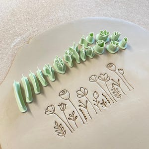 Puede incluir: Un conjunto de sellos de arcilla de plástico verde claro con diversas formas botánicas y florales, dispuestos en fila junto a una pieza plana de arcilla gris claro que ha sido impresa con los diseños de flores y hojas correspondientes.