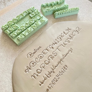 Puede incluir: Un juego de sellos de plástico de color verde claro con letras y números junto a una placa de arcilla gris claro. La arcilla presenta impresiones en relieve del alfabeto en mayúsculas, minúsculas, los números 0-9 y los símbolos # y &.
