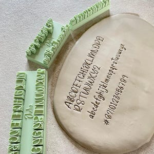 Puede incluir: Tres sellos alfabéticos rectangulares de plástico verde claro con letras, números y símbolos en relieve. Una pieza de arcilla de color beige claro, plana y ovalada, muestra los caracteres impresos por los sellos, incluyendo mayúsculas y minúsculas.