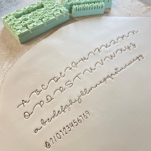 Może przedstawiać: Zestaw jasnozielonych plastikowych stempli z literami i cyframi do odcisków w glinie. Stemple zawierają wielkie i małe litery, cyfry od zera do dziewięciu oraz symbole. Powierzchnia gliny pokazuje pełny odciśnięty alfabet.