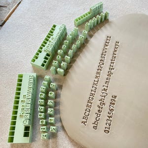 Könnte beinhalten: Ein Set aus hellgrünen Kunststoffstempeln für Buchstaben und Zahlen, angeordnet in Blöcken und Einzelteilen, neben einem flachen Stück hellbraunem Ton mit eingeprägten Großbuchstaben, Kleinbuchstaben und Zahlen von 0 bis 9.