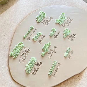 Puede incluir: Una colección de sellos de arcilla de plástico verde claro dispuestos sobre una pieza circular y plana de arcilla de color beige claro. Cada sello presenta una frase alemana única y un icono de corazón, creando impresiones en relieve.