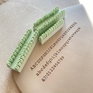 May include: Three pale green rectangular blocks featuring raised alphabet and number stamps. The stamps are positioned next to a flat piece of clay that shows the impressed text: ABCDEFGHIJKLMNOPQRSTUVWXYZ, abcdefghijklmnopqrstuvwxyz, and &?!0123456789.