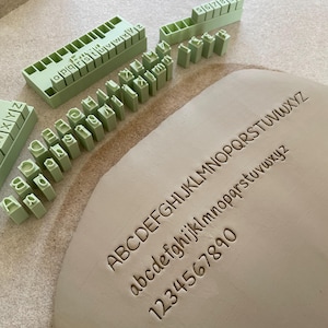 Może przedstawiać: Zestaw jasnozielonych plastikowych stempli z alfabetem i cyframi ułożonych w rzędy. Stemple służą do wyciskania znaków w płaskim kawałku jasnoszarej gliny, na której widnieje wielki alfabet, mały alfabet oraz cyfry 1234567890.