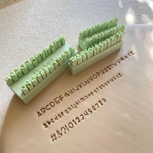 Pode incluir: Um conjunto de carimbos de plástico verde-claro para alfabeto e números dispostos em três blocos. Os carimbos apresentam letras maiúsculas, minúsculas, os números de zero a nove e símbolos como #, &, ?, ! pressionados na argila.