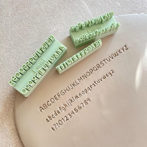 May include: Three light green rectangular clay stamps featuring the brand name Emmi and various letters, numbers, and punctuation marks. These tools are used to press the alphabet, numbers, and symbols into a flat, circular piece of light gray clay.