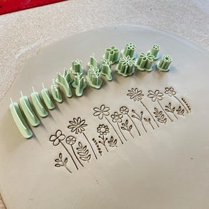 Pode incluir: Um conjunto de carimbos de argila de plástico verde claro com várias formas botânicas e florais. Os carimbos estão dispostos em fila ao lado de uma peça plana de argila que exibe os desenhos impressos de flores, caules e folhas.