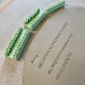 Könnte beinhalten: Ein Set aus hellgrünen, 3D-gedruckten Alphabetstempeln und einem flachen Stück Ton. Die Stempel zeigen Großbuchstaben, Kleinbuchstaben, Zahlen und Symbole. Der Ton zeigt die eingeprägten Texte: Anna, ABCDEFGHIJKLMNOPQRSTUVWXYZ, abcdefghijklmnopqrstuvwxyz und #&?!0123456789.