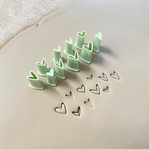 May include: A collection of light green heart-shaped clay cutters arranged in a group, with several heart-shaped impressions stamped into a smooth, flat slab of light beige clay, demonstrating the use of the tools for pottery and crafting.