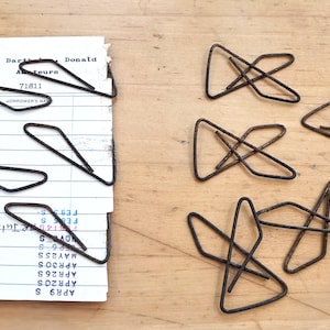 May include: A stack of white library checkout cards with black text including "Bartholomew, Donald" and "Amateurs" held together by three dark, rusted metal wire paper clips, alongside five additional identical rusted wire paper clips.