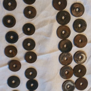May include: A collection of 20 round, metal, antique-looking tokens with a hole in the center. The tokens are arranged in two rows on a white fabric surface.