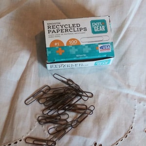 May include: A box of 100 smooth recycled paperclips in a blue and green box with the text "pen+GEAR" and "SMOOTH RECYCLED PAPERCLIPS" on the front. The box is sitting on a white surface with a pile of paperclips scattered around it.