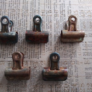May include: Six rusty metal binder clips, some with a blue tint, are arranged on a page of a vintage book. The book page has text printed in black ink.