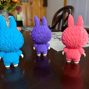 May include: Three colorful bunny figurines in blue, purple, and pink. Each figurine has a textured, furry appearance with upright ears, small arms, and white feet. They are displayed on a dark brown surface.