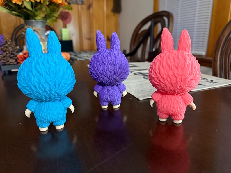 May include: Three colorful bunny figurines in blue, purple, and pink. Each figurine has a textured, furry appearance with upright ears, small arms, and white feet. They are displayed on a dark brown surface.