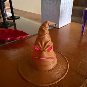 May include: A brown, animated Sorting Hat with glowing red eyes and mouth. The hat sits on a round, textured base. The hat is a decorative item.