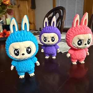 May include: Three colorful figurines of a bunny-like creature with a grumpy expression. The figurines are blue, purple, and pink, with white faces and large, star-shaped eyes. They have fluffy bodies and stand on a dark surface.