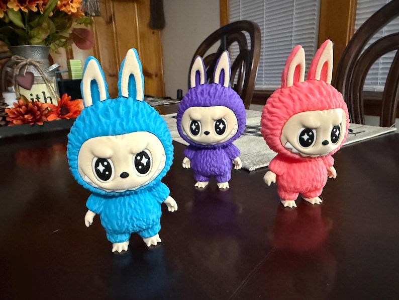 May include: Three colorful figurines of a bunny-like creature with a grumpy expression. The figurines are blue, purple, and pink, with white faces and large, star-shaped eyes. They have fluffy bodies and stand on a dark surface.