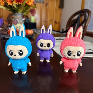 May include: Three colorful figurines of a creature with bunny ears and a round, furry body. The figurines are blue, purple, and pink, each with a white-faced mask and angry eyes. They stand on a dark brown surface.
