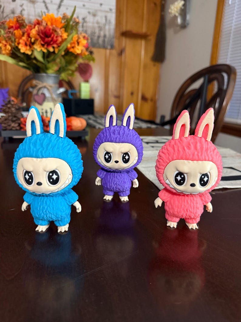 May include: Three colorful figurines of a creature with bunny ears and a round, furry body. The figurines are blue, purple, and pink, each with a white-faced mask and angry eyes. They stand on a dark brown surface.