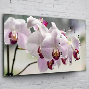 Könnte beinhalten: Eine rechteckige Wanduhr mit einer Nahaufnahme von weißen und lila Orchideenblüten. Das Zifferblatt hat schwarze Zahlen, die die Stunden anzeigen. Der Hintergrund ist ein weiches, verschwommenes Bild eines Fensters.