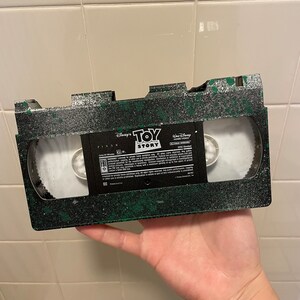 Pu&ograve; includere: Un nastro VHS verde e nero con un'etichetta che recita "Disney Pixar Toy Story" e "Walt Disney Home Video".