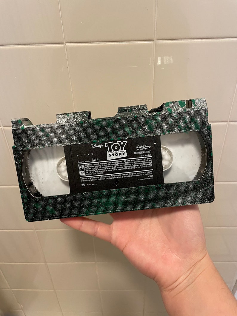 Pu&ograve; includere: Un nastro VHS verde e nero con un'etichetta che recita "Disney Pixar Toy Story" e "Walt Disney Home Video".