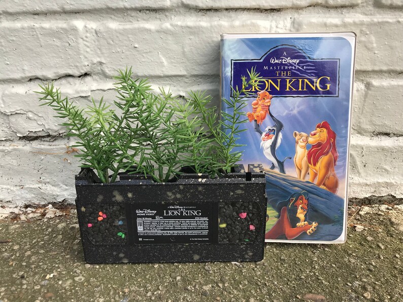 Pu&ograve; includere: Un vaso da fiori nero a forma di cassetta VHS con piante verdi che crescono al suo interno. La cassetta &egrave; decorata con glitter colorati e ha le parole "Il Re Leone" sull'etichetta. Accanto al vaso c'&egrave; una custodia VHS del film "Il Re Leone".