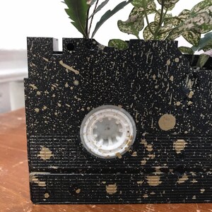 Pu&ograve; includere: Un vaso da fiori nero a forma di cassetta VHS con schizzi di vernice dorata. La cassetta VHS ha un centro in plastica bianca ed &egrave; piena di piante verdi.