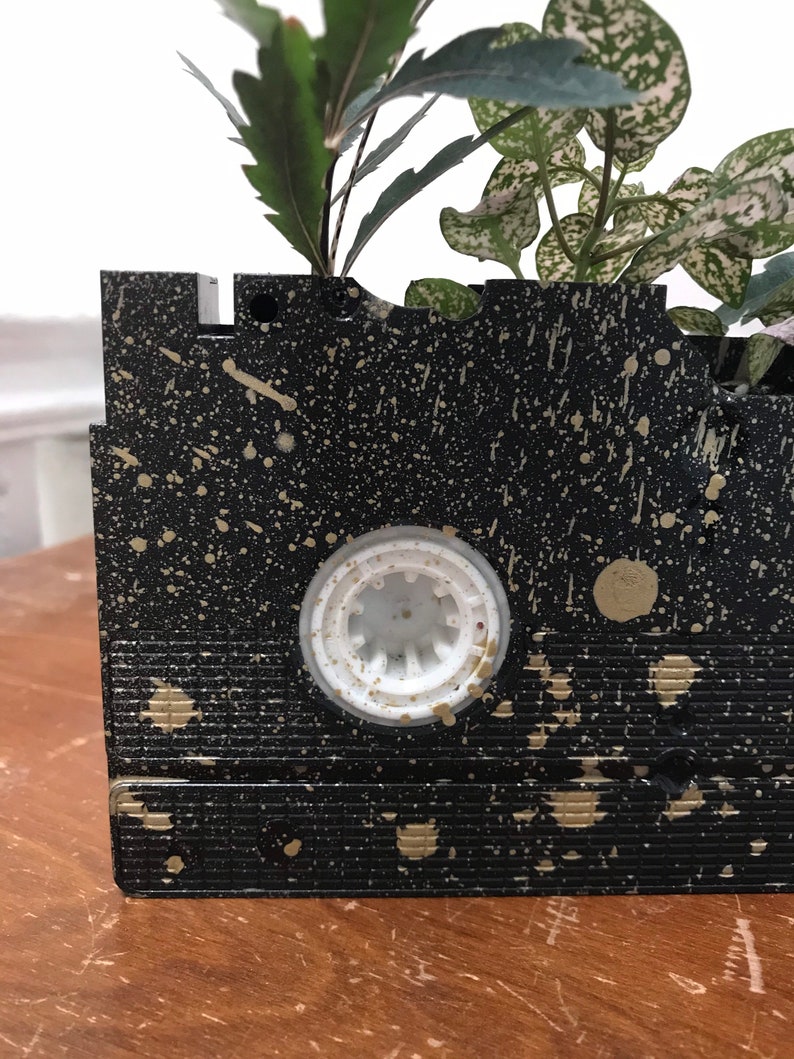 Pu&ograve; includere: Un vaso da fiori nero a forma di cassetta VHS con schizzi di vernice dorata. La cassetta VHS ha un centro in plastica bianca ed &egrave; piena di piante verdi.