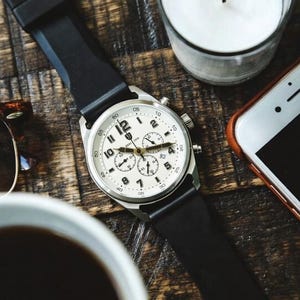 Puede incluir: Un reloj de pulsera con esfera blanca, números negros y correa negra descansa sobre una superficie de madera. El reloj tiene tres esferas más pequeñas y una caja plateada. Otros elementos incluyen un teléfono inteligente, gafas de sol, una vela y una taza de café.