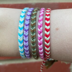 May include: Six colourful friendship bracelets in a variety of shades, including blue, purple, green, pink, and red. Each bracelet has a chevron pattern. The bracelets are worn on a wrist against a brick background.