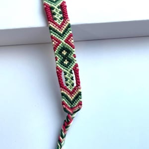 May include: A handmade friendship bracelet with a geometric pattern. The bracelet features a repeating design of red, green, and cream-coloured threads. The bracelet is knotted and braided, with a braided end for tying.