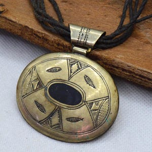 May include: A brass-colored pendant necklace with a black oval center stone and intricate etched designs. The pendant hangs from multiple strands of dark cord. The necklace rests on a wooden surface.