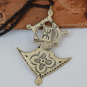 May include: A vintage silver-toned pendant necklace with a black beaded chain. The pendant features an intricate design with a floral pattern and geometric shapes. The necklace is displayed on a textured, brown surface.