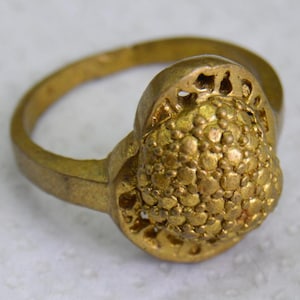 Op de afbeelding: Een goudkleurige ring met een getextureerd, afgerond middengedeelte. De band van de ring is glad, wat leidt tot een gedetailleerd, geclusterd ontwerp op de voorkant. De ring heeft een antieke of vintage uitstraling.