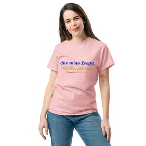 Pode incluir: Uma t-shirt rosa claro com o texto "Che m'ne frega!" em azul e dourado. Abaixo, um desenho de folhas douradas e as palavras "A Neapolitan state of mind" em rosa. A t-shirt está sendo usada.