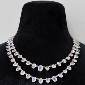 Könnte beinhalten: Doppelreihige Halskette mit schillernden, tropfenförmigen Mondsteinperlen. Die Halskette ist mit kleinen, rosa Perlen aufgereiht, was einen zarten Kontrast erzeugt. Der Schmuck wird auf einer schwarzen Schaufensterpuppe präsentiert.
