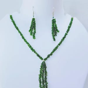 Puede incluir: Un conjunto de joyas de piedras preciosas verdes, que incluye un collar y pendientes a juego, exhibido en un maniquí blanco. El collar presenta una cascada de piedras verdes, y los pendientes tienen un diseño similar.