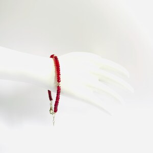 Könnte beinhalten: Ein rotes Perlenarmband mit Silberverschluss wird auf einer weißen Mannequinhand gezeigt. Das Armband besteht aus kleinen, runden, roten Perlen. Der Verschluss ist ein einfacher Haken- und Ösenverschluss.