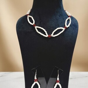 Könnte beinhalten: Ein Schmuckset bestehend aus einer Halskette und einem Paar Ohrhänger. Die Halskette besteht aus weißen perlenartigen Kugeln, die in ovalen Schlaufen angeordnet sind und mit dunkelroten Kugeln akzentuiert werden. Die passenden Ohrhänger spiegeln dieses Design wider.