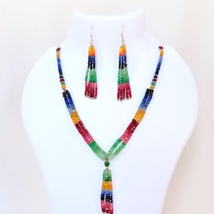Könnte beinhalten: Ein farbenfrohes Perlenketten- und Ohrring-Set. Die Halskette hat eine Kaskade aus leuchtenden Perlen in Rot, Gelb, Grün, Blau und Lila. Die Ohrringe spiegeln das Design der Halskette wider, mit baumelnden Perlenquasten. Der Schmuck wird auf einer weißen Schaufensterpuppe präsentiert.