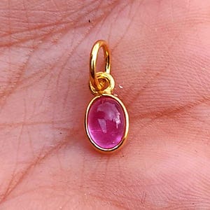 Puede incluir: Un pequeño colgante dorado con una vibrante gema rosa de forma ovalada. El colgante está suspendido de un lazo en la parte superior. La gema está engastada en un bisel dorado. El colgante se muestra sobre un fondo color piel.