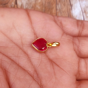 Peut inclure: Un pendentif en forme de cœur avec une pierre précieuse rouge facettée sertie dans un métal doré. Le pendentif est petit et délicat, avec une bélière simple pour une chaîne. La pierre a une couleur rouge vive.
