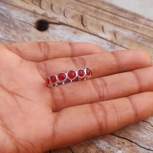 Può includere: Un anello a fascia color argento caratterizzato da una fila continua di piccole pietre cabochon rotonde di un rosso vibrante, incastonate in una montatura metallica. L'anello presenta una finitura lucida e un design uniforme con ogni pietra.