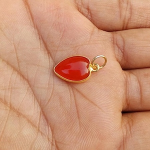 May include: A small heart-shaped pendant featuring a smooth, vibrant red stone set within a polished gold-toned metal frame. The pendant includes a circular gold-toned jump ring at the top, designed for attachment to a necklace chain or bracelet.