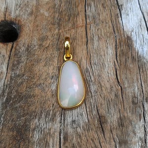 Puede incluir: Un colgante de oro con una piedra preciosa blanca, iridiscente, en forma de pera. La piedra preciosa está engastada en un bisel de oro y tiene una superficie facetada. El colgante cuelga de una fianza de oro. El fondo es una superficie de madera envejecida.